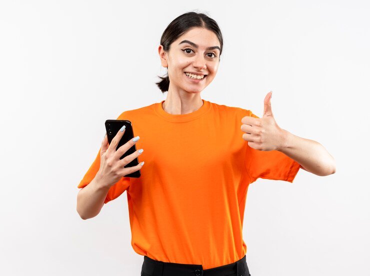 young girl wearing orange t-shirt holding smartphone showing thumbs up smiling cheerfully standing white wall